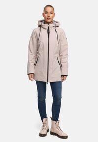 Lichtroze waterdichte jas met een capuchon, voorzijde met rits, zwarte accenten en dubbele zijzakken. Draagt bij donkere jeans en beige veterschoenen.