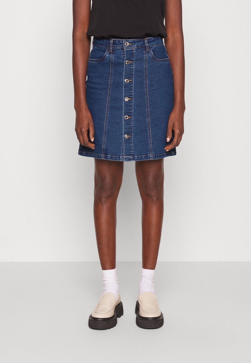 Femme portant une jupe en jean bleue à boutons sur le devant, un haut noir, des chaussettes blanches et des mocassins beiges à semelles épaisses, debout devant un fond uni.