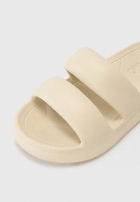 Beige slide sandal with two wide straps, made of textured rubber material. The sole is flat with a rounded edge and smooth inner surface.
