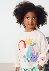 Sudadera color crema con diseños bordados coloridos, que incluyen un arcoíris, sol, flor y formas de animales en la parte delantera y la manga.