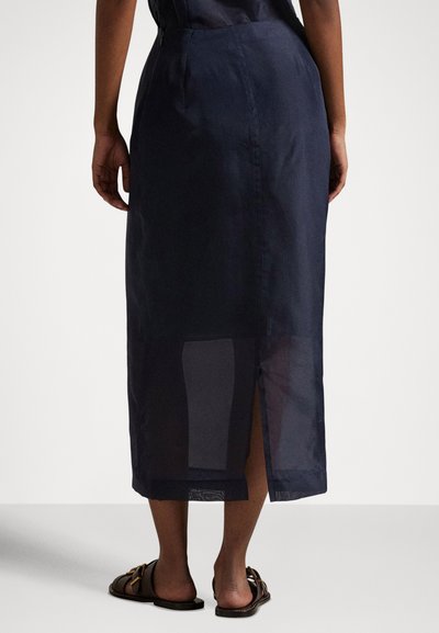 Skats no muguras uz personu, kas valkā tumši zilu midi svārku ar atvērumu aizmugurē un tumši brūnas plakanas sandales uz vienkārša fona.