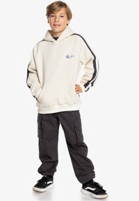 Felpa crema con cappuccio e tasca frontale, maniche a righe nere e bianche, con un logo a forma di montagna blu; abbinata a pantaloni cargo scuri.