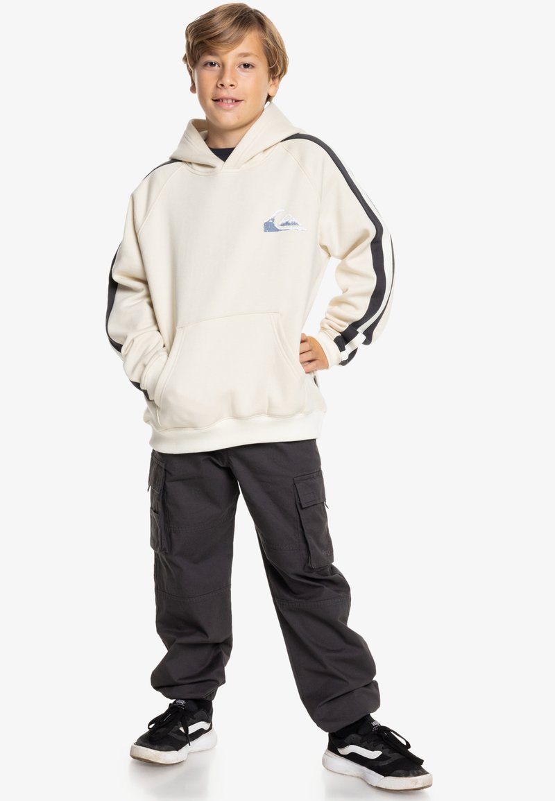 Felpa crema con cappuccio e tasca frontale, maniche a righe nere e bianche, con un logo a forma di montagna blu; abbinata a pantaloni cargo scuri.