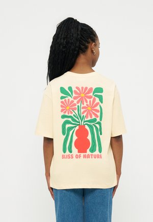 Άτομο με πλεξούδες που φοράει ανοιχτό μπεζ μπλουζάκι με κόκκινο βάζο με ροζ λουλούδια και πράσινα φύλλα, με το κείμενο «Bliss of Nature» από κάτω.