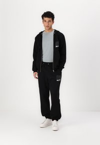 Sort zip-up hoodie over en lys grå T-shirt og sorte sweatpants med lommer. Hvide sneakers. Minimalistisk design med diskret branding.