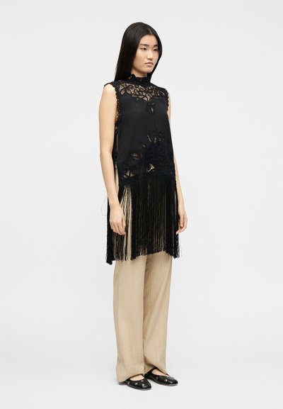 Γυναίκα με μακριά μαύρα μαλλιά που φοράει μαύρο δαντελένιο τοπ χωρίς μανίκια με κρόσσια πάνω από μπεζ φαρδιά παντελόνια και μαύρα επίπεδα παπούτσια.