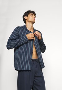 Camicia a righe blu navy con colletto, maniche lunghe e una vestibilità comoda. Abbinata a pantaloni scuri e larghi. Tessuto dalla consistenza liscia.