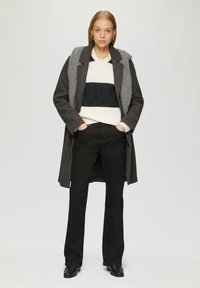 Grijze lange jas met een gestructureerde grijs en zwarte capuchon, gedragen over een crème trui met zwarte strepen, gecombineerd met zwarte uitlopende broek en laarzen.