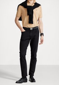 Camisa polo color beige, jeans negros, suéter negro de punto drapeado sobre los hombros, cinturón negro y zapatos de cuero negros. Diseño ajustado y minimalista.