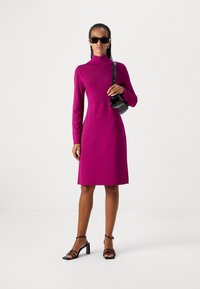 Fuchsia langärmeliges Kleid mit hochgeschlossenem Kragen, gerippter Textur und schmaler Passform. Dazu getragen werden schwarze Sonnenbrillen und eine kleine Handtasche, kombiniert mit Riemchen-Absätzen.