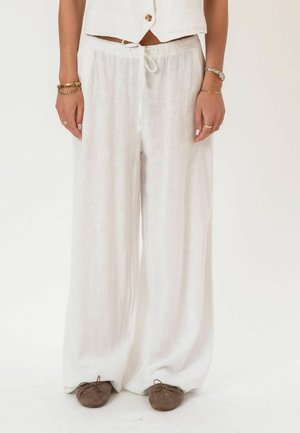 Nő, aki laza fehér vászonnadrágot visel, bokánál megkötött bézs balerinacipőkkel, kezén karkötők és gyűrűk díszítik, egyszerű fehér háttér előtt áll.