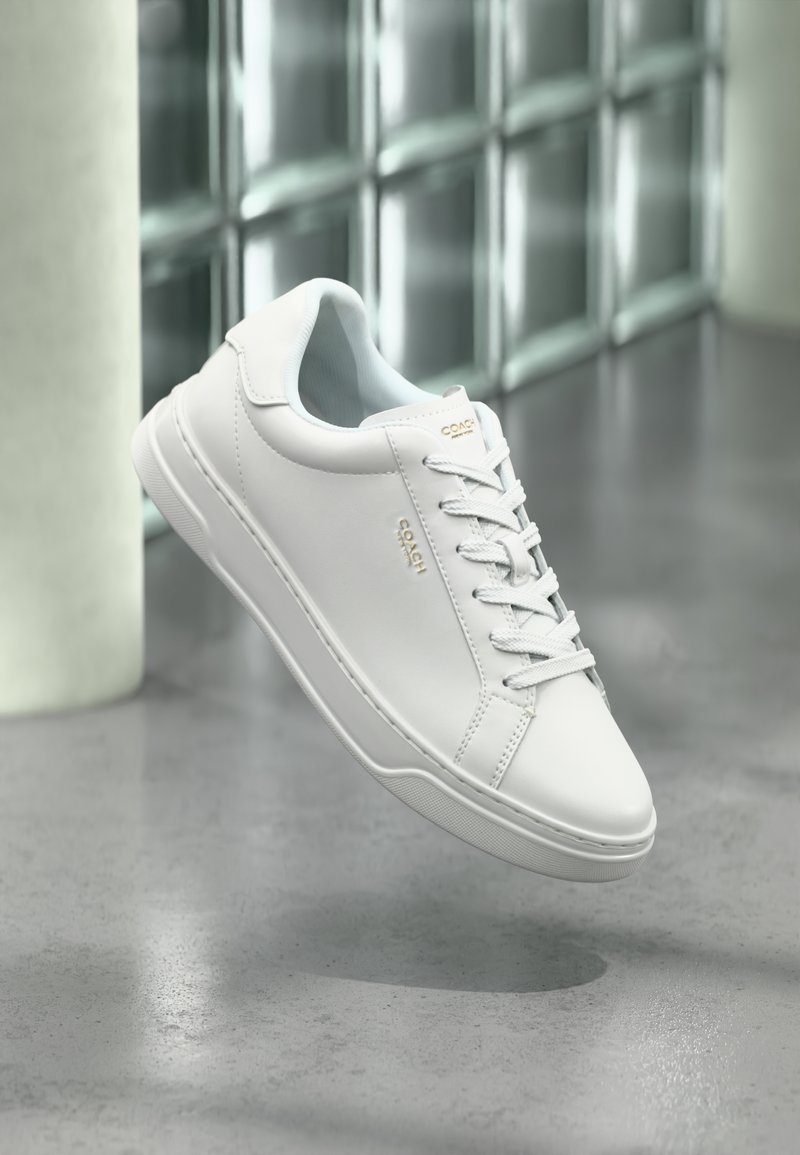 Baskets en cuir blanc de la marque Coach avec lacets, flottant au-dessus d'un sol en béton poli près d'un mur en blocs de verre et d'une colonne dans un cadre intérieur moderne.