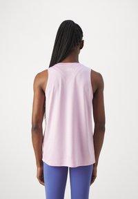 Kvinna med långt flätat hår som bär en ljusrosa ärmlös topp och blå leggings, sedd bakifrån mot en enfärgad bakgrund.