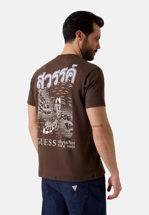 Uomo che indossa una maglietta marrone con disegno di farfalla bianca e testo thailandese sul retro, abbinata a jeans blu scuro.