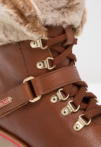 Bruna läderstövlar med en krage av konstfäll, prydda med guldfärgad hårdvara, bruna snören med röda accenter och en remdetalj på sidan.