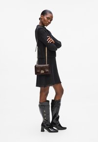Schwarzer Strickpullover, schwarzes fransiges Kleid, glänzende schwarze knielange Stiefel mit Knöpfen und eine dunkelbraune Handtasche aus Lackleder mit einer Kettenriemen.