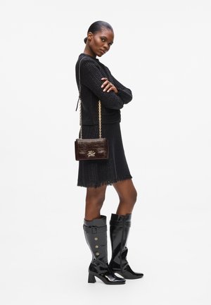 Schwarzer Strickpullover, schwarzes fransiges Kleid, glänzende schwarze knielange Stiefel mit Knöpfen und eine dunkelbraune Handtasche aus Lackleder mit einer Kettenriemen.