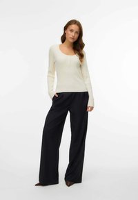 Maglia a maniche lunghe a coste color crema con scollo a barchetta e dettagli con bottoni, abbinata a pantaloni neri a vita alta a gamba larga.