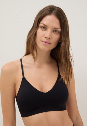 Femme aux longs cheveux bruns portant un bralette noir à fines bretelles, face à l'avant sur un fond clair uni.