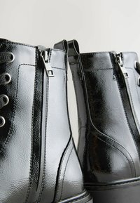 Botas de tobillo de imitación de cuero negro con acabado brillante, que presentan ojales de metal, cremalleras y detalles texturizados a lo largo de los costados.