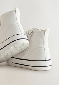 Witte canvas enkellaarzen met een hoge bovenkant, een zijrits en een zwarte streep langs de zool. Textuur rubberen buitenzool voor grip.