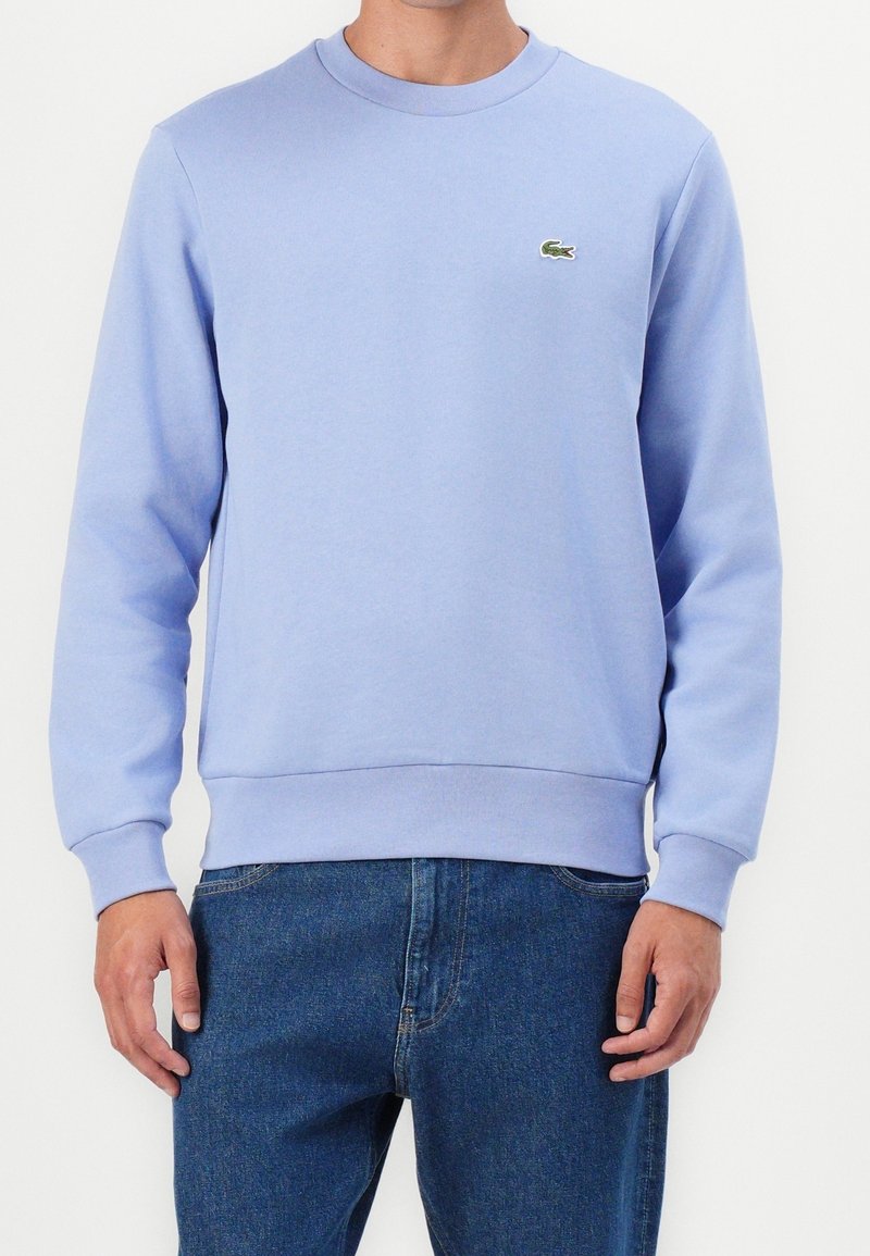 Homme portant un sweat-shirt bleu clair à col rond avec un petit logo crocodile vert sur la poitrine et un jean bleu.