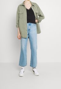 Camicia di cotone khaki con bottoni sopra una canotta nera, abbinata a jeans svasati azzurri chiari e sneakers bianche con accenti neri.