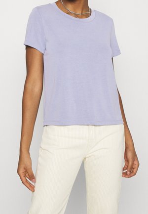 Persona con una camiseta de manga corta de color púrpura claro y pantalones de color crema con sutiles rayas verticales, de pie frente a un fondo liso.