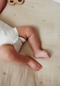 Body alb acoperă un picior și un picioruș dolofan pe o pătură bej texturată. Un jucărie de lemn cu accente albastre este vizibilă în apropiere.