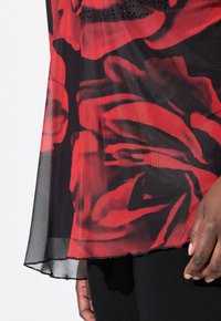 Top nero trasparente con grande motivo a rosa rossa, caratterizzato da un orlo arrotondato e un bordo leggermente sfilacciato. I dettagli includono piccoli accenti neri sul collo.