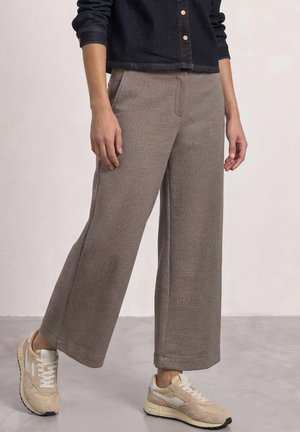 Personne portant un pantalon marron à jambes larges, des baskets beige et une chemise en denim foncé, debout sur un sol gris.