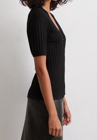 Top de punto negro con textura de canalé, mangas cortas, escote en V y seis botones frontales; combinado con una falda de cuero lisa y oscura.