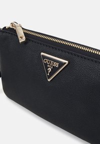 Černá texturovaná syntetická kabelka s zlatým zipem a trojúhelníkovou plaketou s logem "GUESS" a znakem "?". Kompaktní design, minimalistický vzhled.