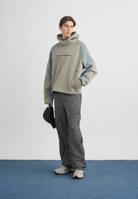 Ljusgrå hoodie med blå detaljer och svart logotyp, kombinerad med mörka cargobyxor. Outfiten har en avslappnad passform och funktionell design.