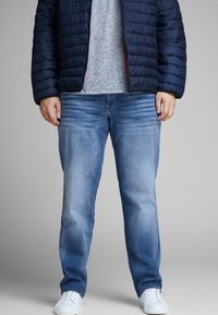 Light blue denim jeans feature a relaxed fit with subtle whiskering, paired with a gray t-shirt and a navy puffer jacket. White sneakers complete the look.