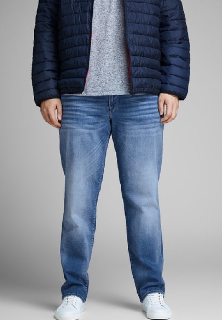 Light blue denim jeans feature a relaxed fit with subtle whiskering, paired with a gray t-shirt and a navy puffer jacket. White sneakers complete the look.