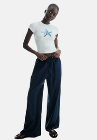 Bela skrajšana majica z grafiko modre morske zvezde, v kombinaciji s prostornimi mornarsko modrimi hlačami. Model nosi črne sandale; ozadje je enostavno belo.