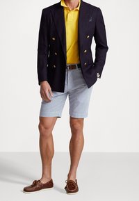 Marinblå dubbelknäppt blazer, gul polo, randiga shorts och bruna seglarskor. Jackan har guldknappar; klocka på handleden.