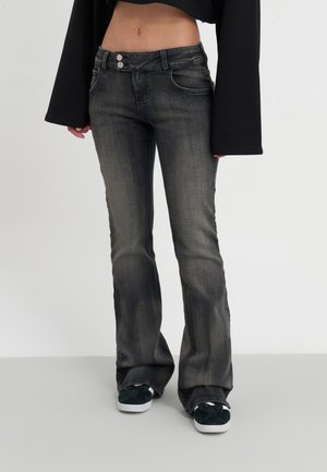 Femme portant un jean noir délavé évasé, un top court noir à larges manches et des baskets noires à semelles blanches, debout devant un fond blanc.