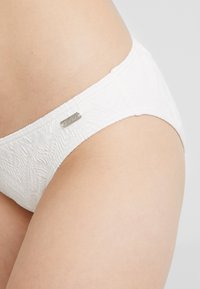 Slip bikini bianco con un motivo testurizzato, caratterizzato da un tessuto liscio, una cintura elastica e un piccolo tag metallico del brand sul lato.