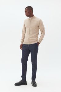 Bēšas krāsas rāvējslēdzēja džemperis ar augstu apkakli, uzvilkts virs tumši ziliem biksēm. Džemperim ir gluda tekstūra un pieguļošs izskats, kopā ar melniem apaviem.