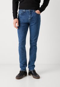 Jeans de mezclilla de lavado oscuro con corte ajustado, que cuentan con cierre de cremallera, diseño de cinco bolsillos y costuras sutiles. Llevados con zapatillas negras.