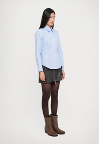 Kvinde står op, iført en lys blå knap-shirt, sort læder mini-nederdel med frontlynlås, sorte strømpebukser og brune ankelstøvler.
