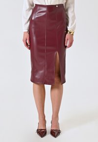 Jupe crayon bordeaux, longueur genoux, en faux cuir brillant avec fente sur le devant, détails cousus et bouton décoratif sur la taille.