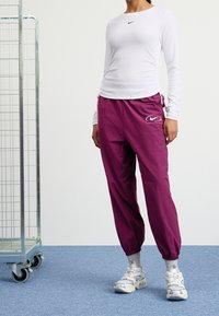 Person som bär en vit långärmad topp och lila Nike-joggingbyxor med vita och grå sneakers, stående på en blå matta bredvid ett metallställ.