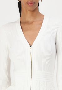 Robe tricotée blanche à rayures avec un décolleté en V profond et des détails de fermeture éclair dorés. Présente un motif texturé en bas et une silhouette cintrée.
