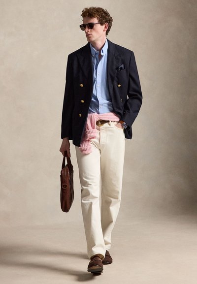 Homme marchant portant des lunettes de soleil, blazer bleu marine avec des boutons dorés, chemise bleu clair, pantalon crème, pull rose noué à la taille, tenant un sac en cuir marron.
