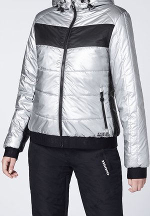 Chaqueta de puff plateada con detalles en negro, cierre con cremallera y capucha. Presenta puños elásticos y un logo en el dobladillo. Diseño ajustado.