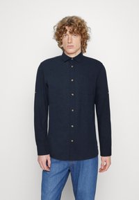 Camicia a maniche lunghe blu navy con bottoni, realizzata in tessuto testurizzato, con colletto, tasca frontale e bottoni marroni. Indossata con jeans blu.