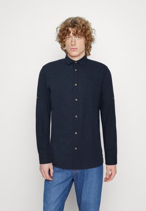 Camicia a maniche lunghe blu navy con bottoni, realizzata in tessuto testurizzato, con colletto, tasca frontale e bottoni marroni. Indossata con jeans blu.